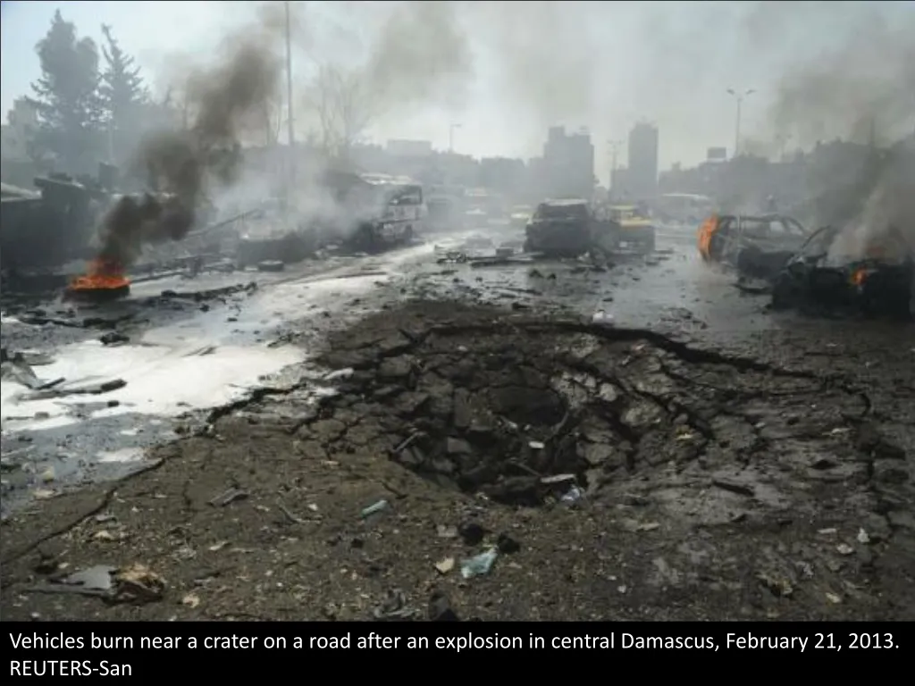 vehicles burn near a crater on a road after